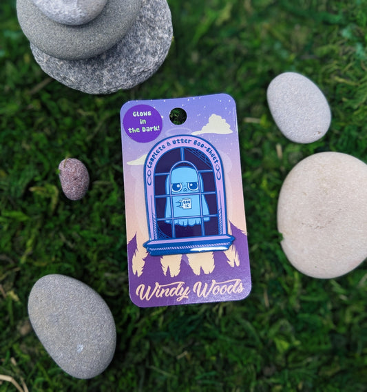 Photograph of a mossy background with an enamel pin of a ghost in a window with a coffee cup. The pin has blue plating and purple enamel. The pin says: “Complete and utter boo-sheet.”