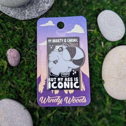 A photograph of an enamel pin on a backing card surrounded by moss and stones. The enamel pin is a rectangle with a raccoon standing with a big butt. The pin says, "My Anxiety is chronic but my ass is iconic."