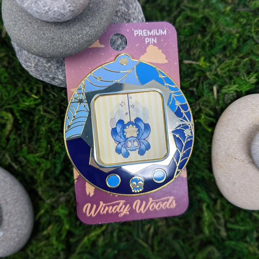 Photograph of a mossy background with an enamel pin of a blue and yellow star spider. The spider is in a tamagotchi type device that is blue with gold webs. The screen of the tamagotchi is lenticular and is animated to show Cosmo climbing down a web.