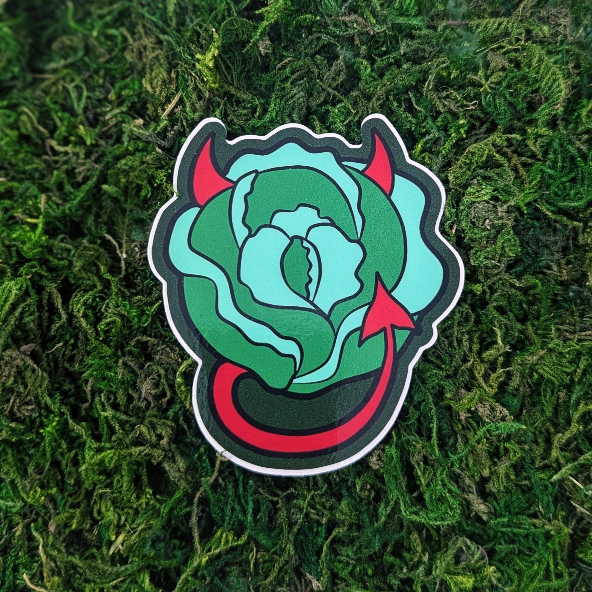 Photograph of a mossy background with a vinyl sticker of a head of lettuce with red horns and a tail.