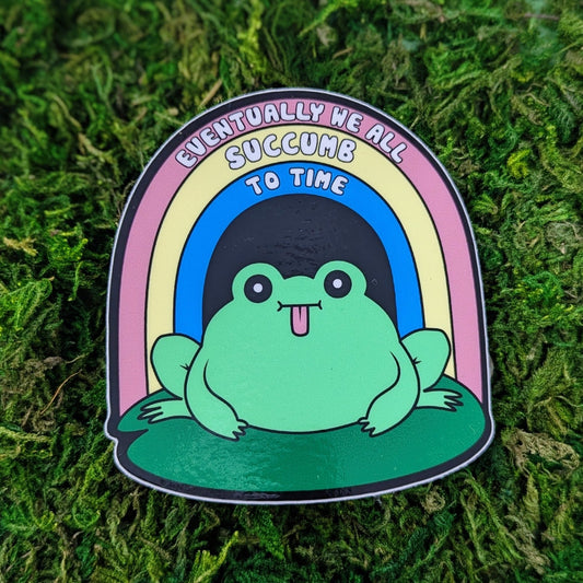 Photograph of a mossy background with a vinyl sticker of a frog on a lily pad with a rainbow over it that says “Eventually we all succumb to time.”