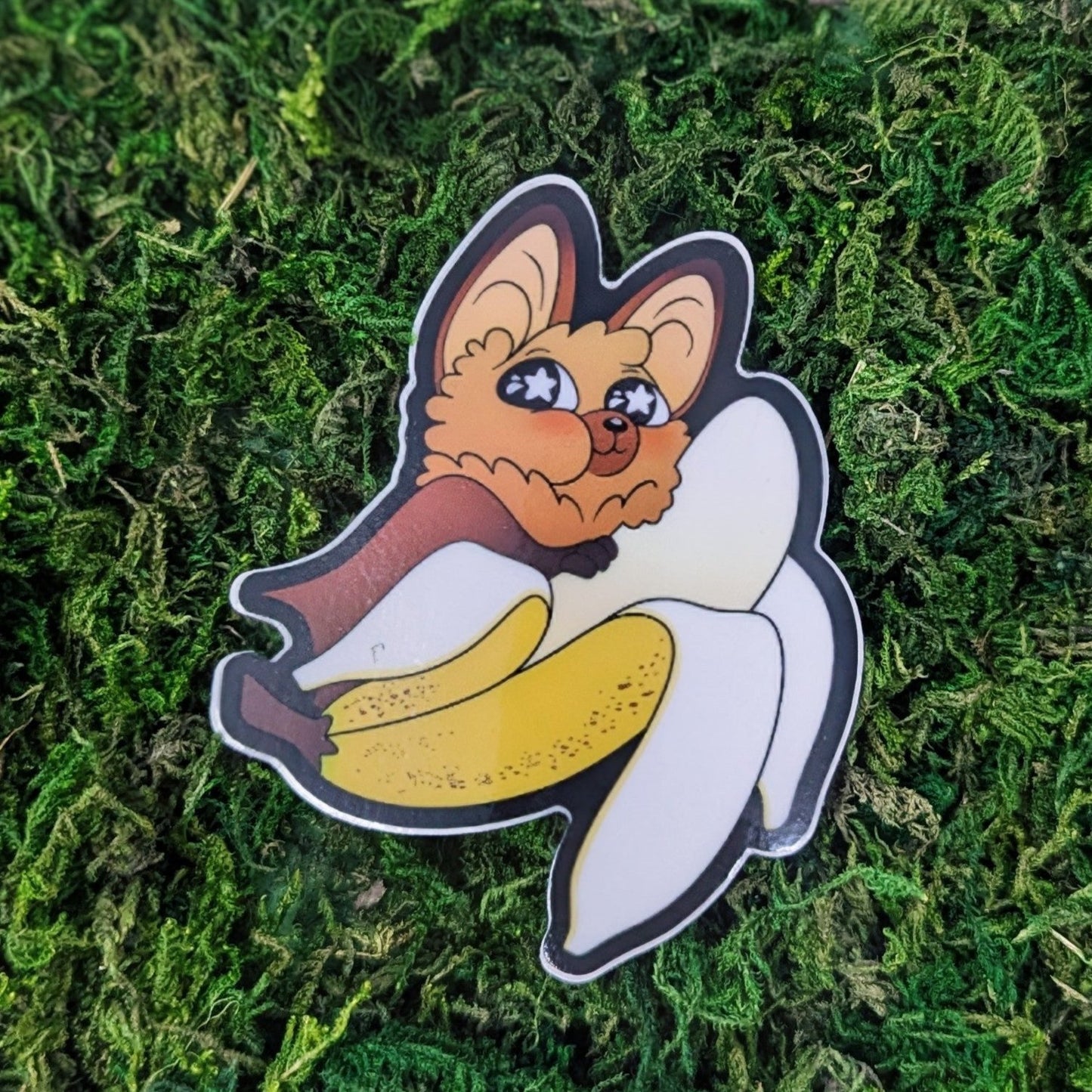 Photograph of a mossy background with a vinyl sticker of a fox faced fruit bat eating a banana.