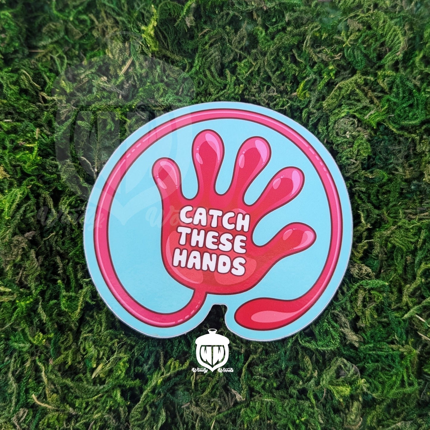 A photograph of a sticker on some moss. The sticker features artwork of a a red slap hand toy on a light blue background. In the middle of the hand it says, "Catch these hands."