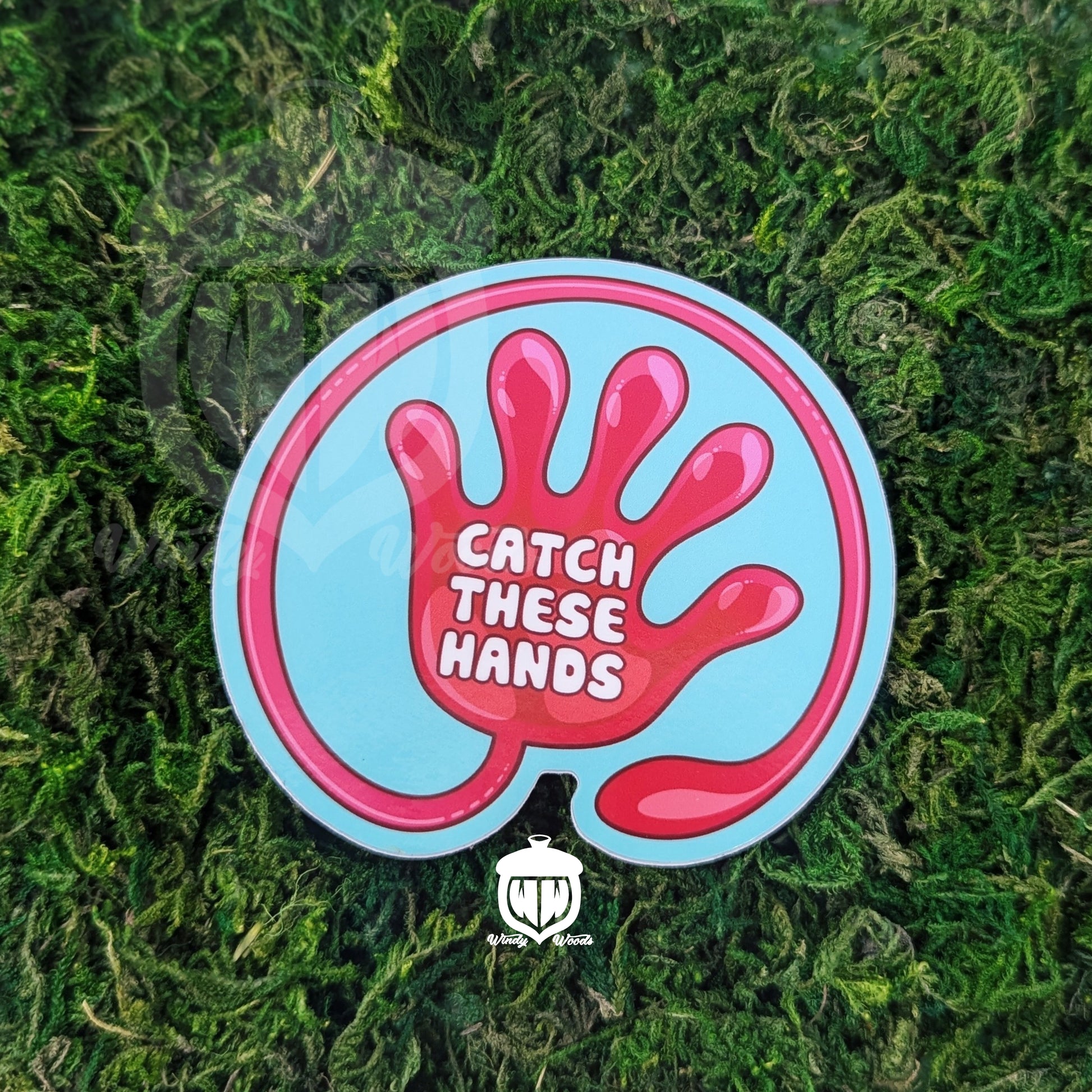 A photograph of a sticker on some moss. The sticker features artwork of a a red slap hand toy on a light blue background. In the middle of the hand it says, "Catch these hands."
