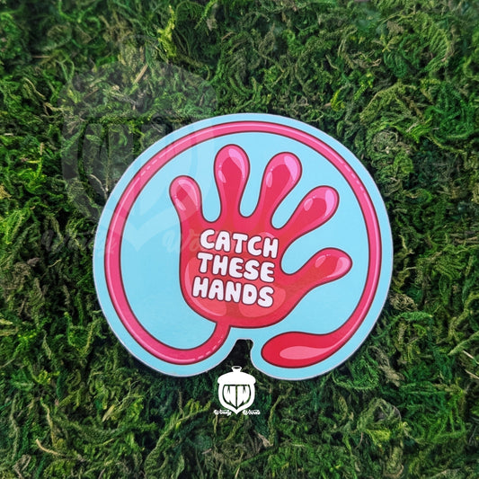 A photograph of a sticker on some moss. The sticker features artwork of a a red slap hand toy on a light blue background. In the middle of the hand it says, "Catch these hands."