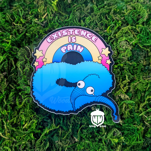 A Photograph of a sticker on some moss. The sticker features a blue toy worm on a string with a sparkly rainbow over it that says, "Existence is Pain."