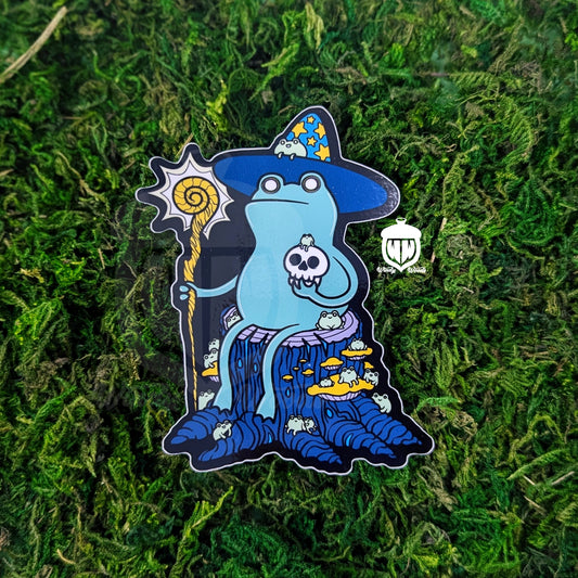A Photograph of a sticker on some moss. The sticker features artwork of a bipedal green frog sitting on a stump. This is the Loveland Frogman Cryptid. The Frogman is wearing a blue wizard hat with yellow stars on it. They're holding a yellow branch staff that's glowing with power. The Frogman is holding a skull in their other hand. They are covered and surrounded by small frogs that they definitely summoned to their side.