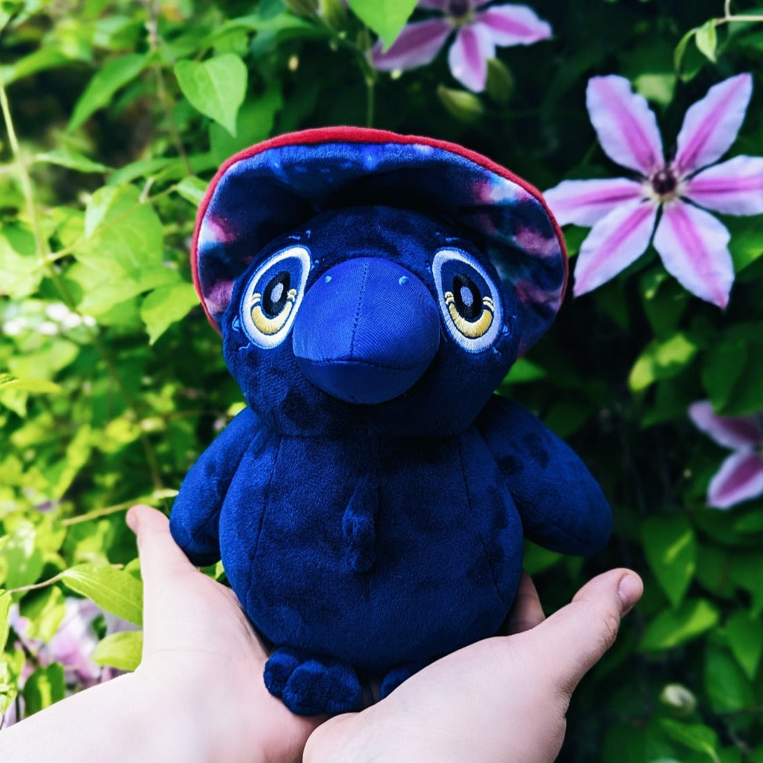 A photograph of someone holding Ikarus the raven witch plush in front of some clematis plants.