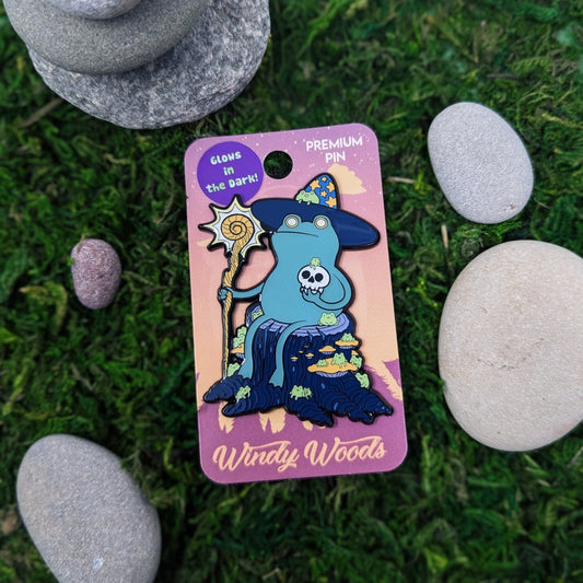 A photograph of an enamel pin on a backing card sitting in moss and stone. The enamel pin is of a bipedal frog sitting on a stump with a wizard hat and glowing staff. The frog is holding a skull and surrounded by little frogs.