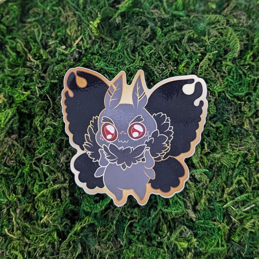 Photograph of a mossy background with a vinyl sticker of Mothman with a gold outline.