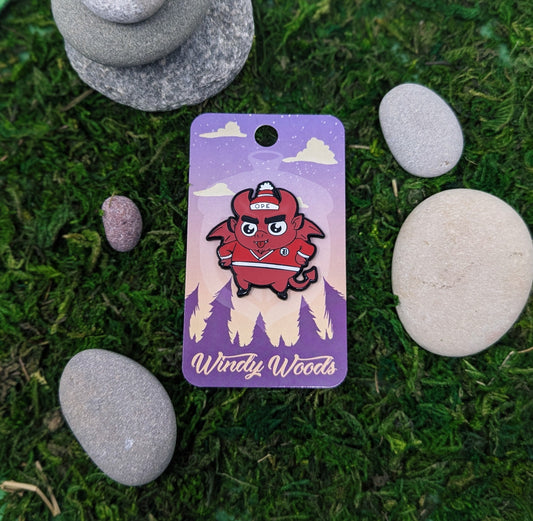 A photograph of an enamel pin on a backing card surrounded by moss and stone. The enamel pin features a little red imp called the Nain Rouge. The Nain Rouge is wearing a red jersey and red winter hat that says, "Ope."