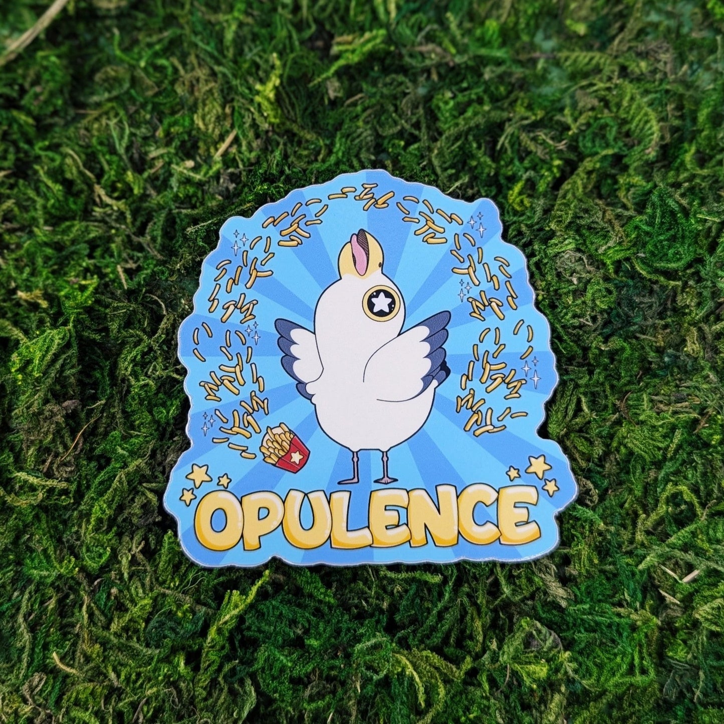Photograph of a mossy background with a vinyl sticker of a seagull surrounded by french fries on a blue background. Under the gull it says, “Opulence.”