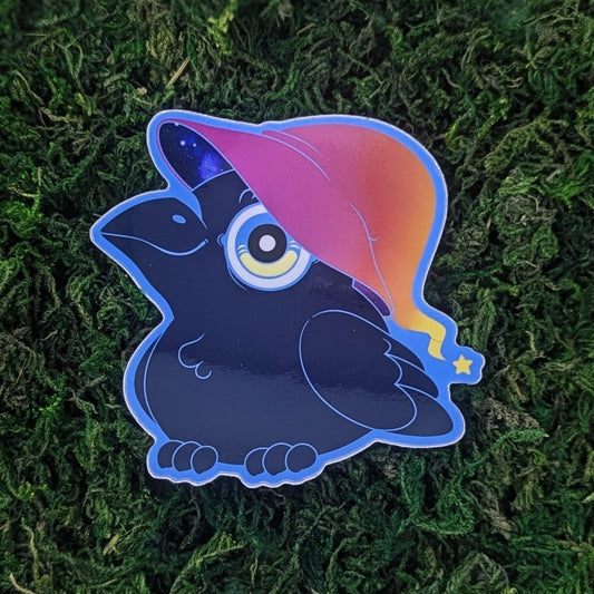 Photograph of a mossy background with a vinyl sticker of a little raven with a sunset gradient witch hat with a star on the end.