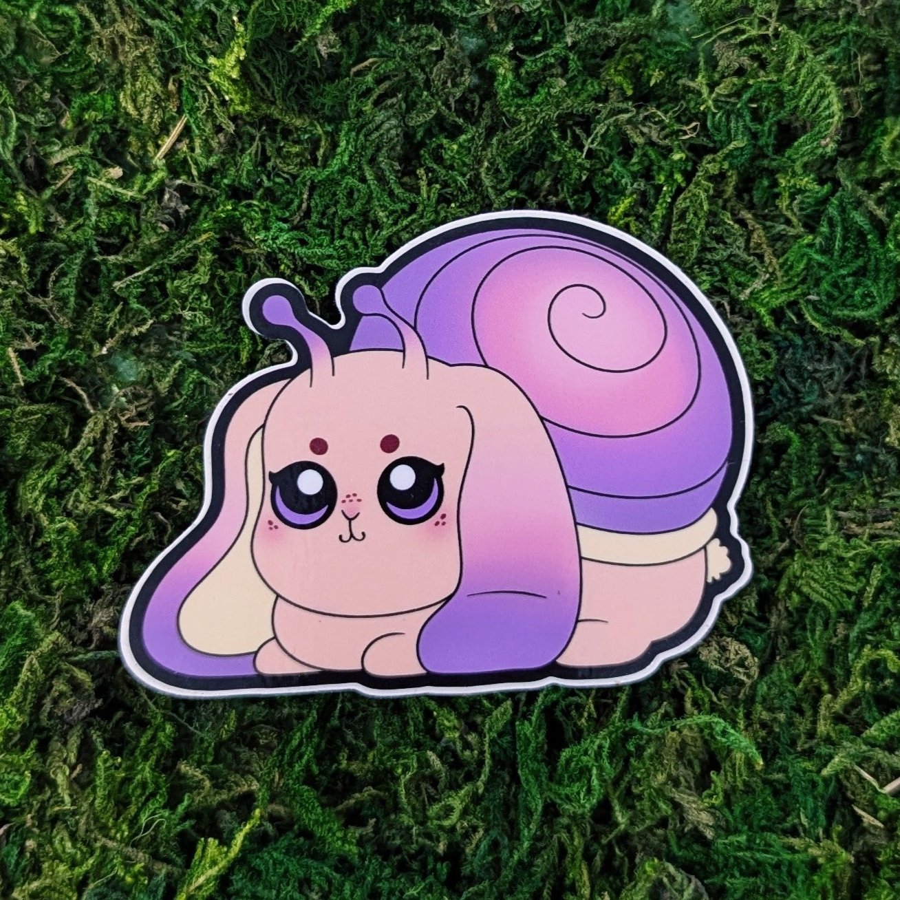 Photograph of a mossy background with a vinyl sticker of a Bunny/Snail hybrid animal called Slobun.