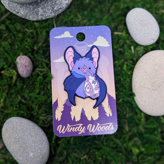 A photograph of an enamel pin on a backing card surrounded by moss and stone. The enamel pin features a purple vampire bat holding a white and pink type O+ juice box.