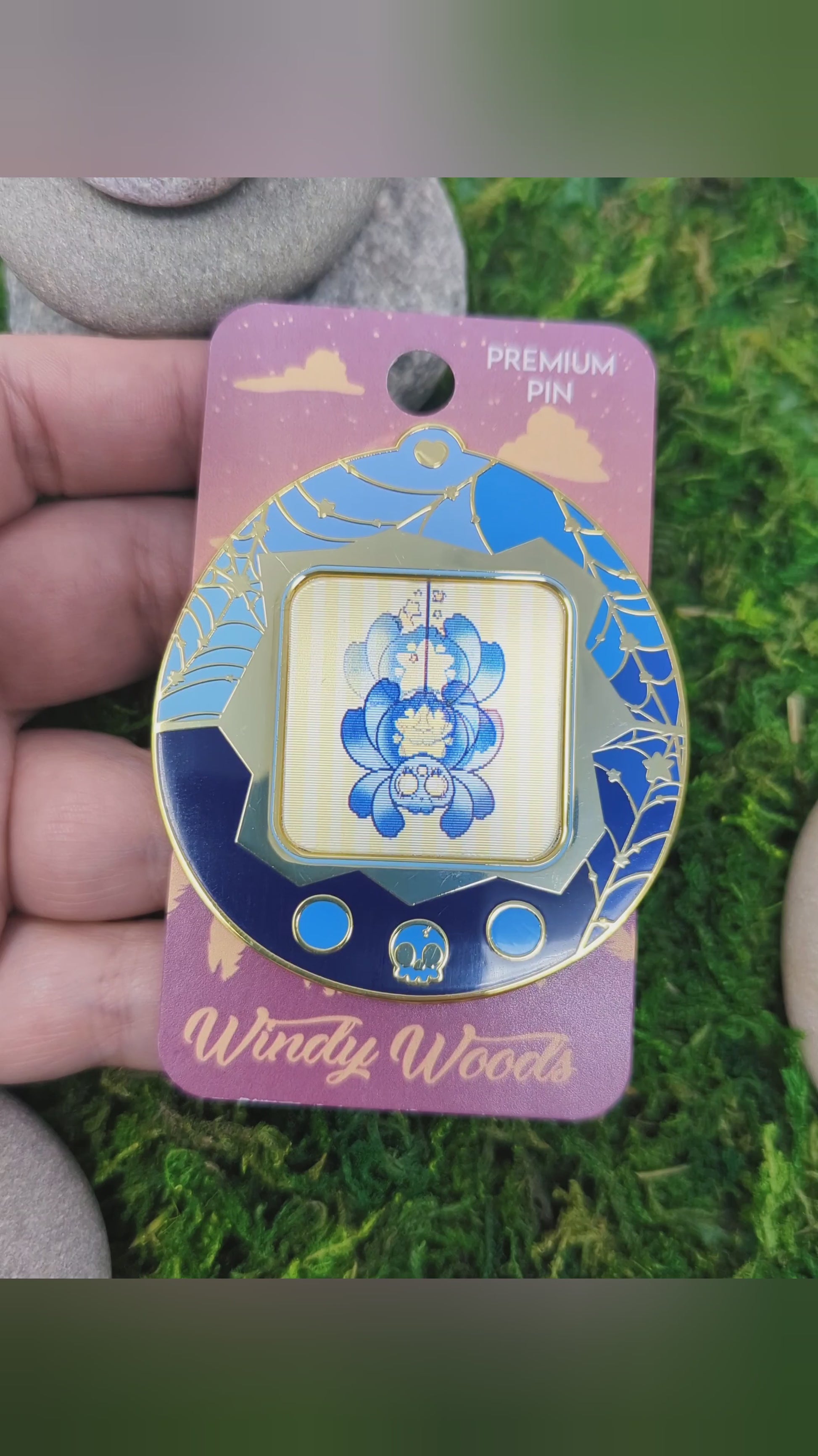 Video of a mossy background with an enamel pin of a blue and yellow star spider. The spider is in a tamagotchi type device that is blue with gold webs. The screen of the tamagotchi is lenticular and is animated to show Cosmo climbing down a web.