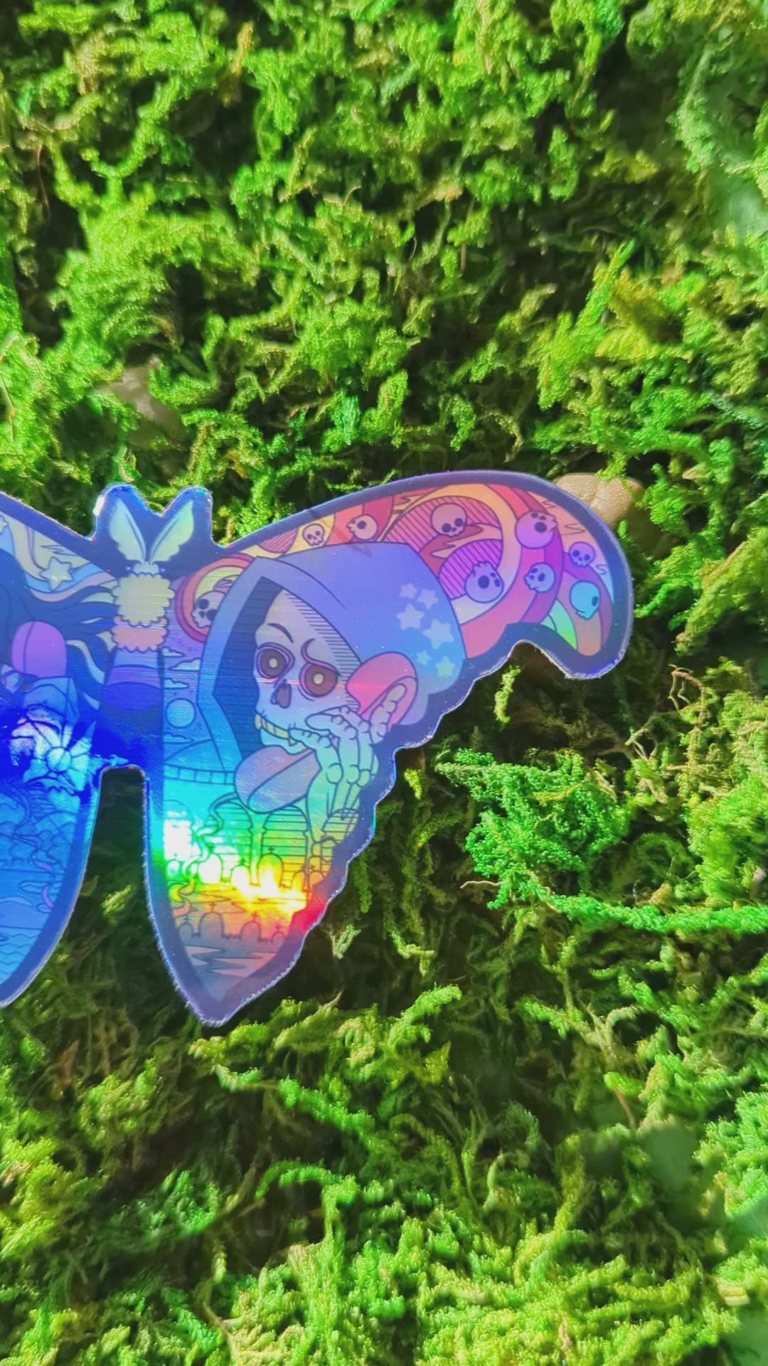 Video of a mossy background with a holographic vinyl sticker of a moth with life and death having a phone chat on the wings. The video shows how shiny the holographic effect is.