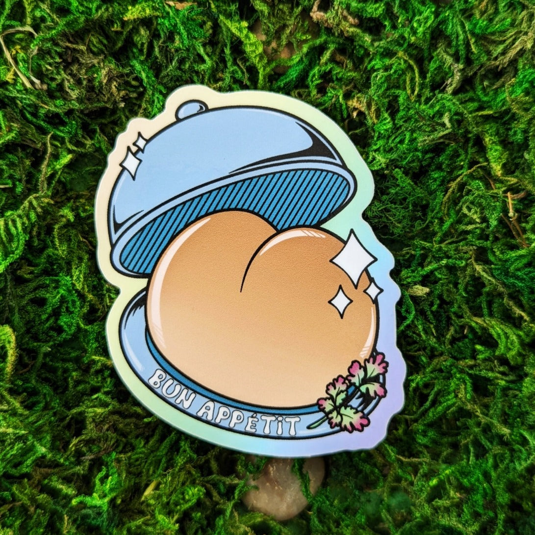 Photograph of a mossy background with a vinyl sticker of a serving tray presenting a booty shaped bread bun. On the bottom plate it says, “Bun Appétit."