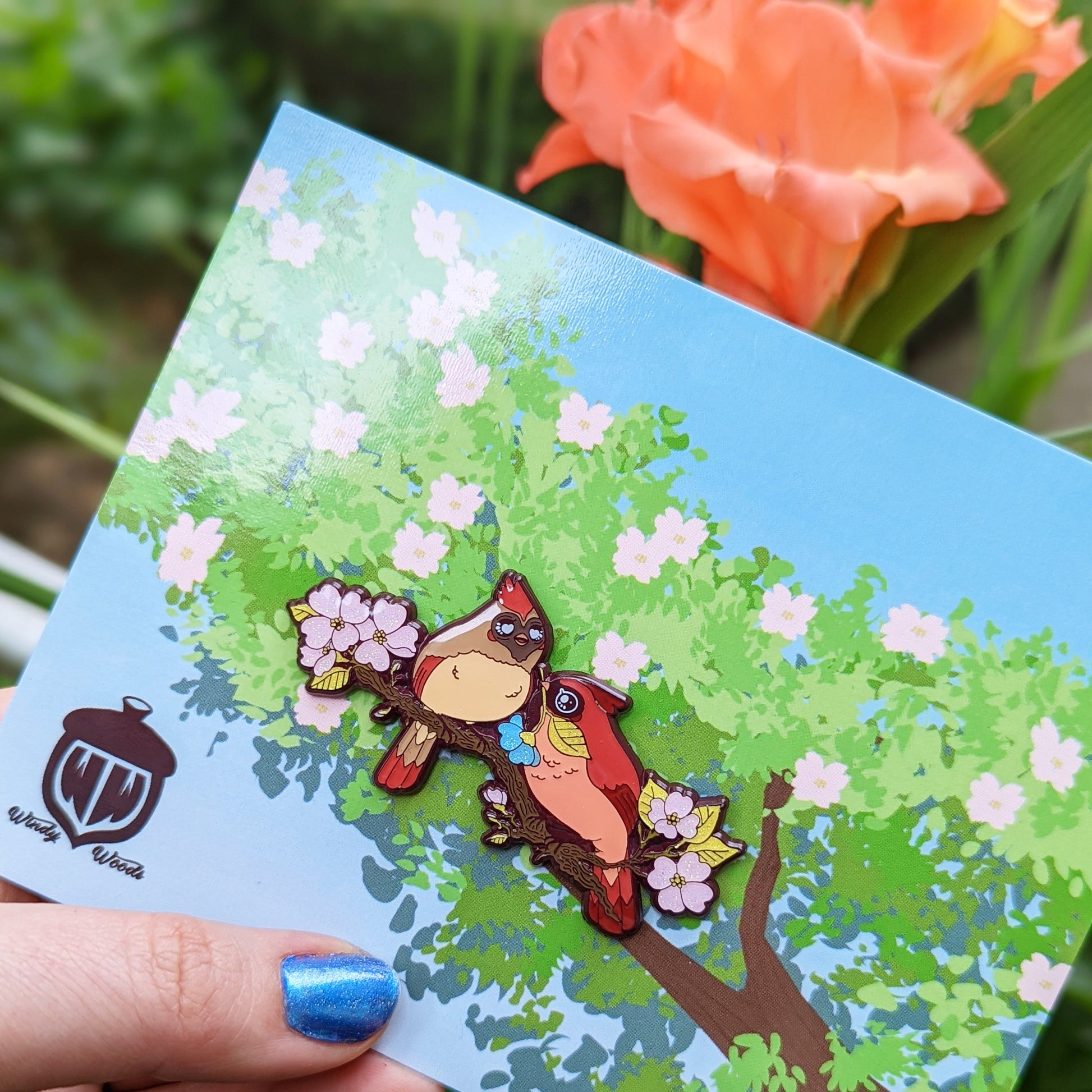 A photograph of a person holding an enamel pin in front of an orange flower. The enamel pin shows two cardinals on a dogwood tree surrounded by pink flowers. One of the cardinals is handing the other a special blue flower.