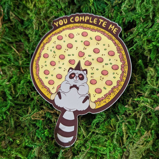 Photograph of a mossy background with a vinyl sticker of a raccoon sitting amongst a pizza in the spot of a missing slice. The raccoon has hearts in his eyes and above the pizza it says, “You complete me.”