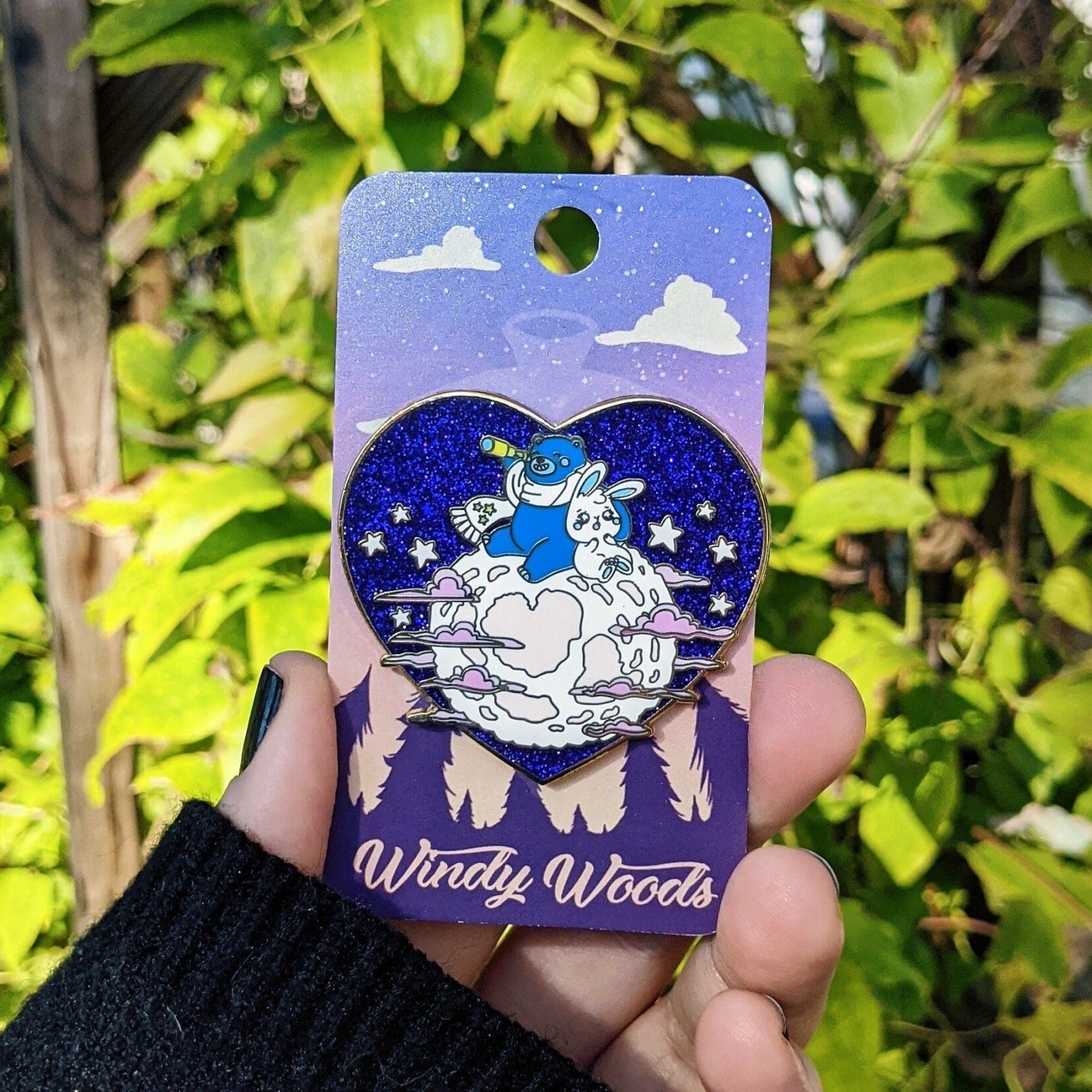A photograph of an enamel pin. The enamel pin is a heart shape. The moon has a blue bear and a white bunny looking at stars.