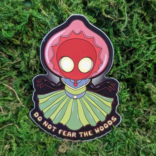 Photograph of a mossy background with a vinyl sticker of Braxxie the Flatwoods Monster. Braxxie appears to have a green dress, red tie, red head, and glowing yellow eyes. Under them it says, “Do not fear the woods.”
