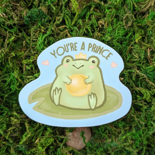 Photograph of a mossy background with a 3 inch vinyl sticker of a Frog holding a gold ball on a lily pad while wearing a crown. Over the frog it says “You’re a Prince.”