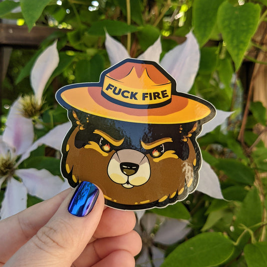 A photograph of a vinyl sticker being held in front of a clematis plant. The sticker is of Smoky the Bear with the reflection of flames in his eyes. His ranger hat says, “Fuck fire.”