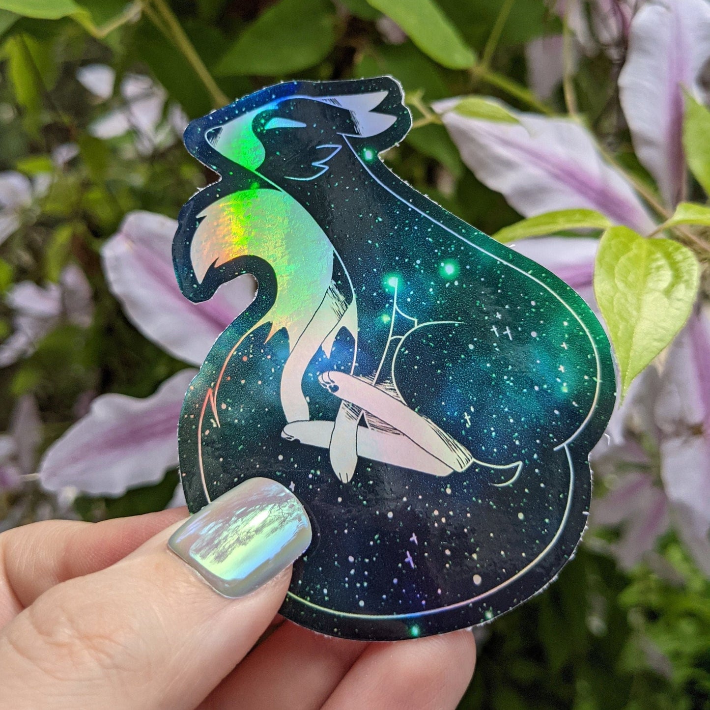 A photograph of a vinyl sticker being held in front of a clematis plant. The vinyl sticker is of a green holographic galaxy themed fox.