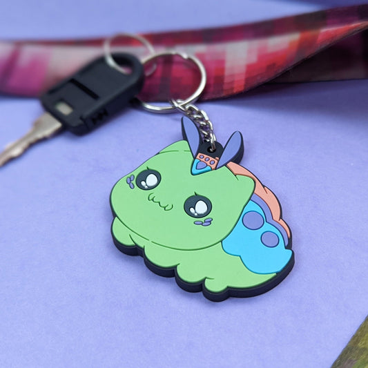 A photograph of a purple background with a landyard and a key. Attached to the key is a PVC Keychain of a cat caterpillar hybrid creature called a Kittypillar.