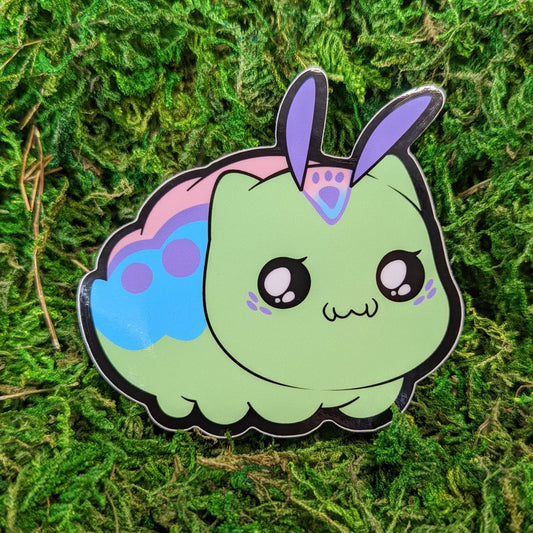 Photograph of a mossy background with a vinyl sticker of a kitten and caterpillar hybrid called Kittypillar.