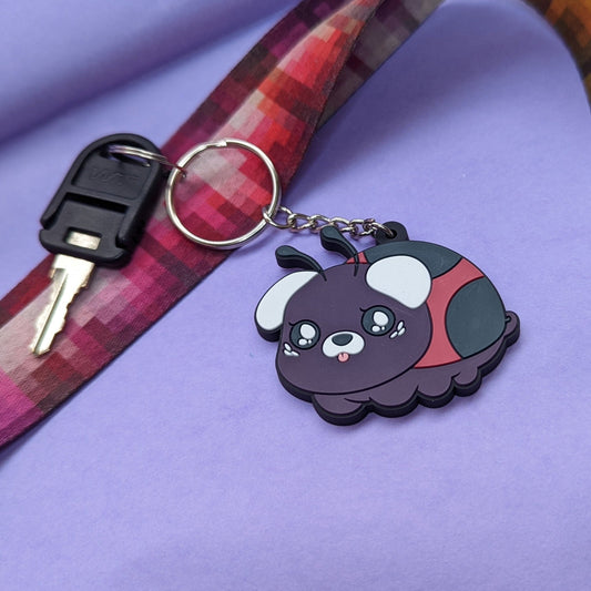 A photograph of a purple background with a landyard and a key. Attached to the key is a PVC Keychain of a pug ladybug hybrid creature called a Ladypug.