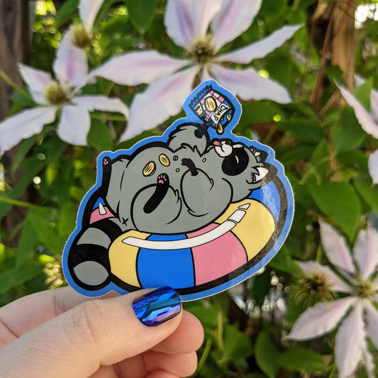 A photograph of a vinyl sticker being held in front of a clematis plant. The vinyl sticker is a raccoon in a floaty while eating chips.