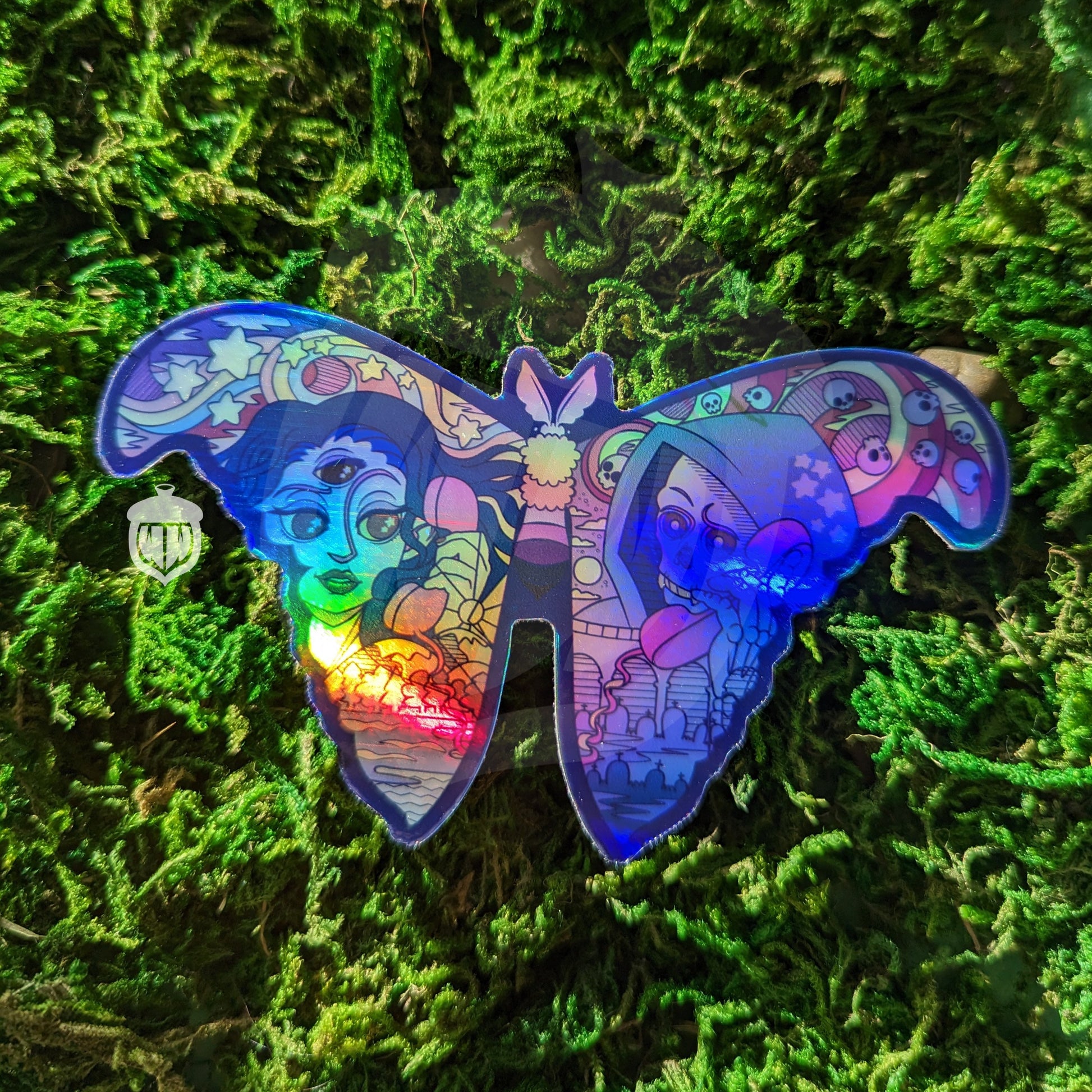 A photograph of a vinyl sticker being held in front of a clematis plant. The vinyl sticker is of a holographic moth. One wing has the representation of life and one has death and they’re both speaking on the phone.