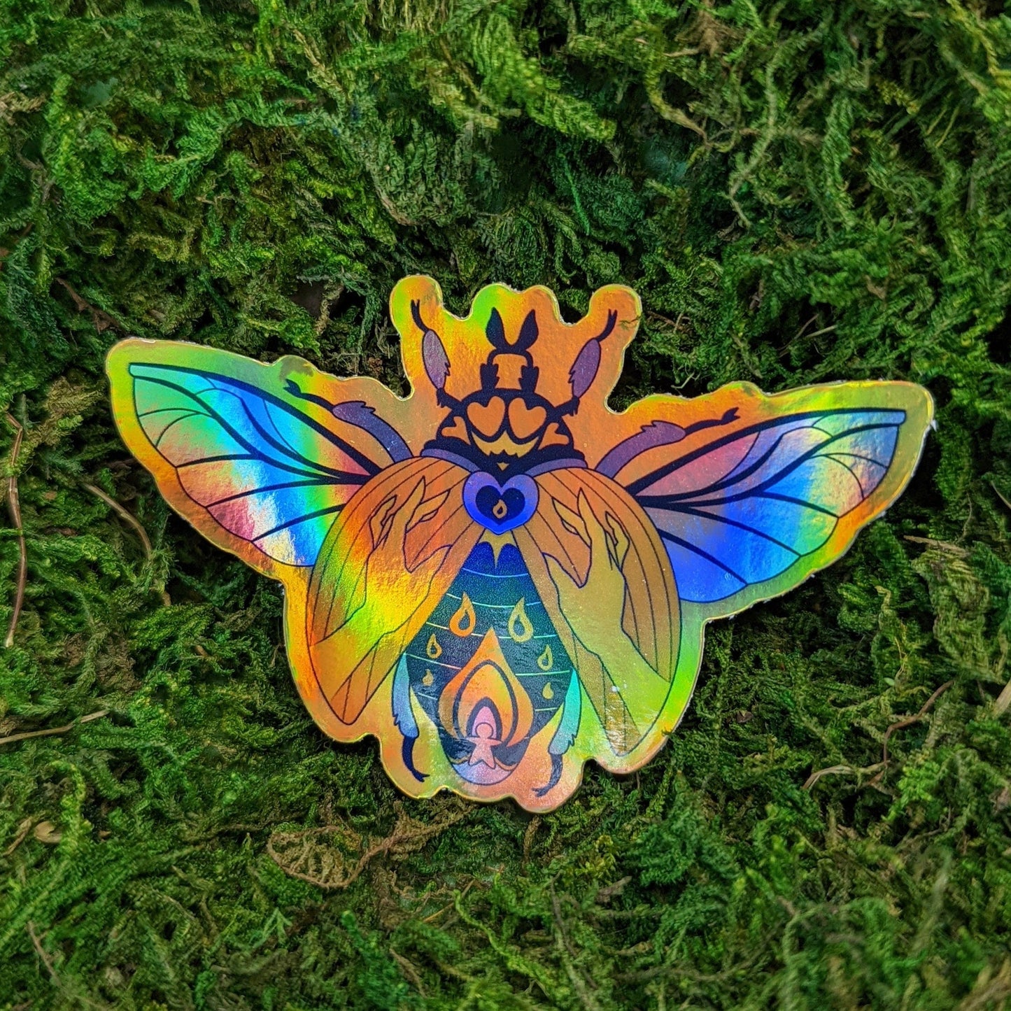 Photograph of a mossy background with a vinyl holographic sticker a gold and blue beetle with hands, heart, and flame designs on its back.