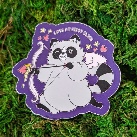 Photograph of a mossy background with a vinyl sticker of a raccoon with a cupid’s bow and arrow and wings. Through the tip of the arrow is a slice of pizza. Above the raccoon it says, “Love at first slice.”