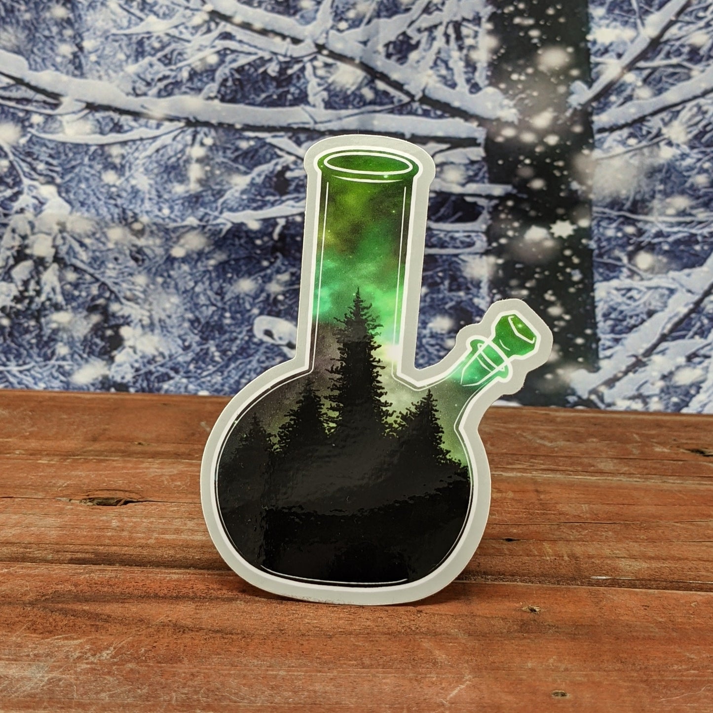 A redwood background with a snowy backdrop. On the wood is a vinyl sticker of a bong with a the northern lights in a boreal forest.