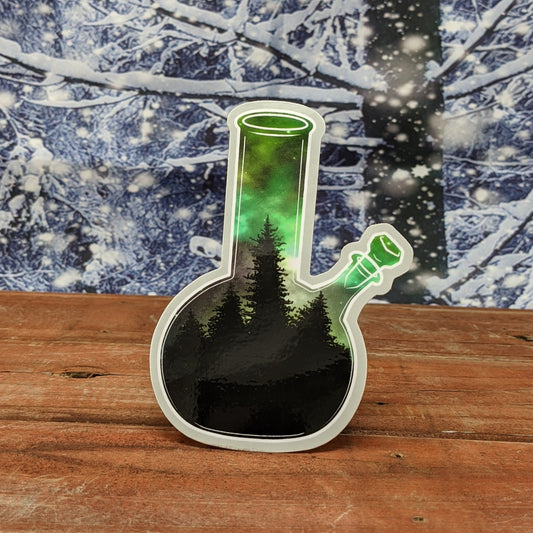 A redwood background with a snowy backdrop. On the wood is a vinyl sticker of a bong with a the northern lights in a boreal forest.