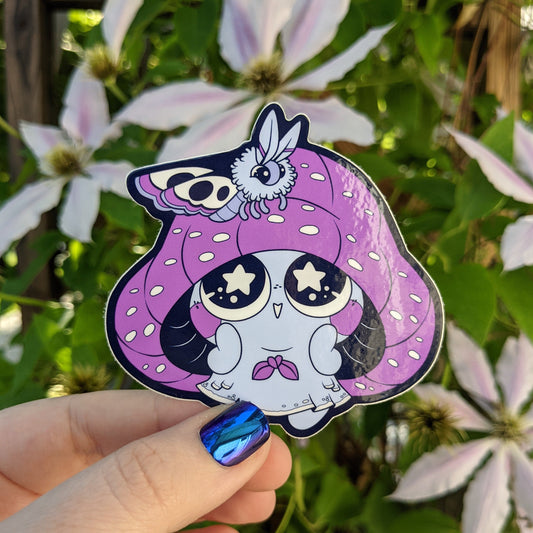 A photograph of a vinyl sticker being held in front of a clematis plant. The sticker is of a purple mushroom sprite called a Mushkin. Mushkin is admiring the moth that landed on its cap.