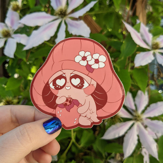 A photograph of a vinyl sticker being held in front of a clematis plant. The sticker is of a red mushroom sprite called a Mushkin. Mushkin is holding a strawberry with a ladybug on it.
