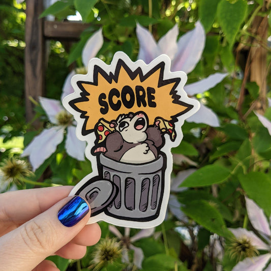 A photograph of a vinyl sticker being held in front of a clematis plant. The vinyl sticker is a opossum coming out of a trash can with two slices of pizza and exclaiming, “SCORE!”