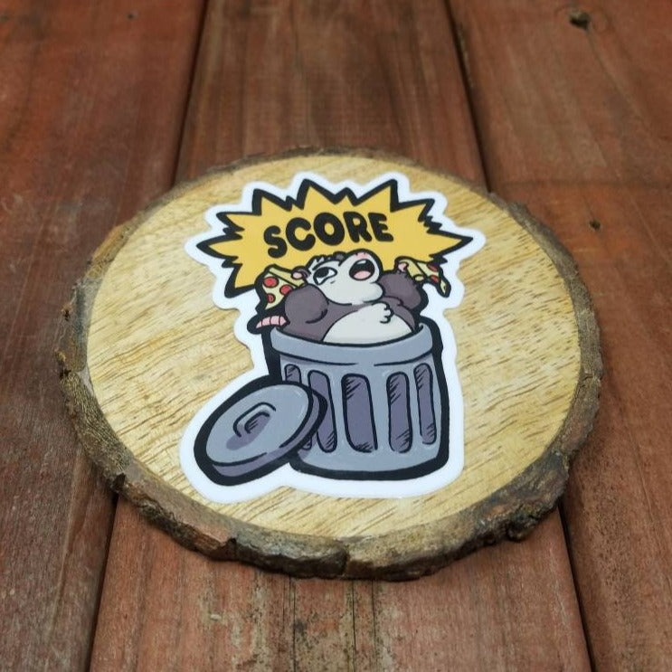 A redwood background with a small wood round. On the wood is a vinyl sticker of a opossum coming out of a trash can with two slices of pizza and exclaiming, “SCORE!”