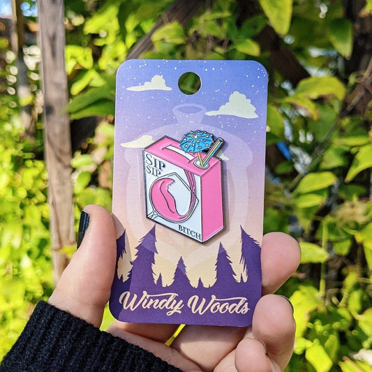 A photograph of an enamel pin being held in front of a clematis plant. The enamel pin is of a box of wine with a drink umbrella coming out the top with a straw. The box has a glass on white on it and it reads, “Sip Sip Bitch.”