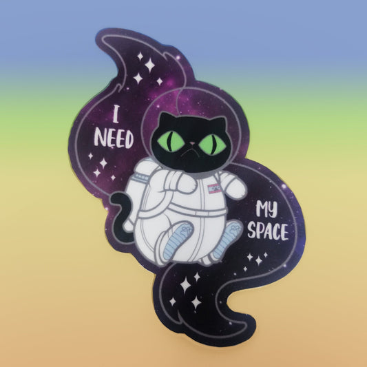 A photograph of a vinyl sticker resting on a pastel rainbow background. The vinyl sticker is of a black cat in a space suit floating through space. The sticker reads, “I need my space.”