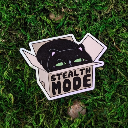 Photograph of a mossy background with a vinyl sticker of a black cat looking smug in a box. The outside of the box says, “Stealth Mode.”