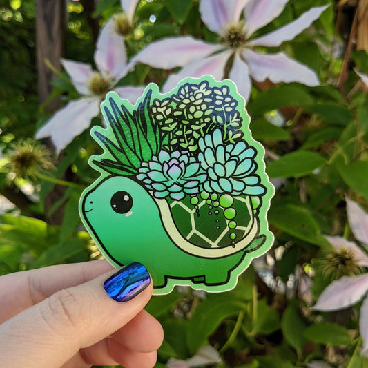 A photograph of a vinyl sticker being held in front of a clematis plant. The vinyl sticker of a green turtle with succulents growing out of its shell.