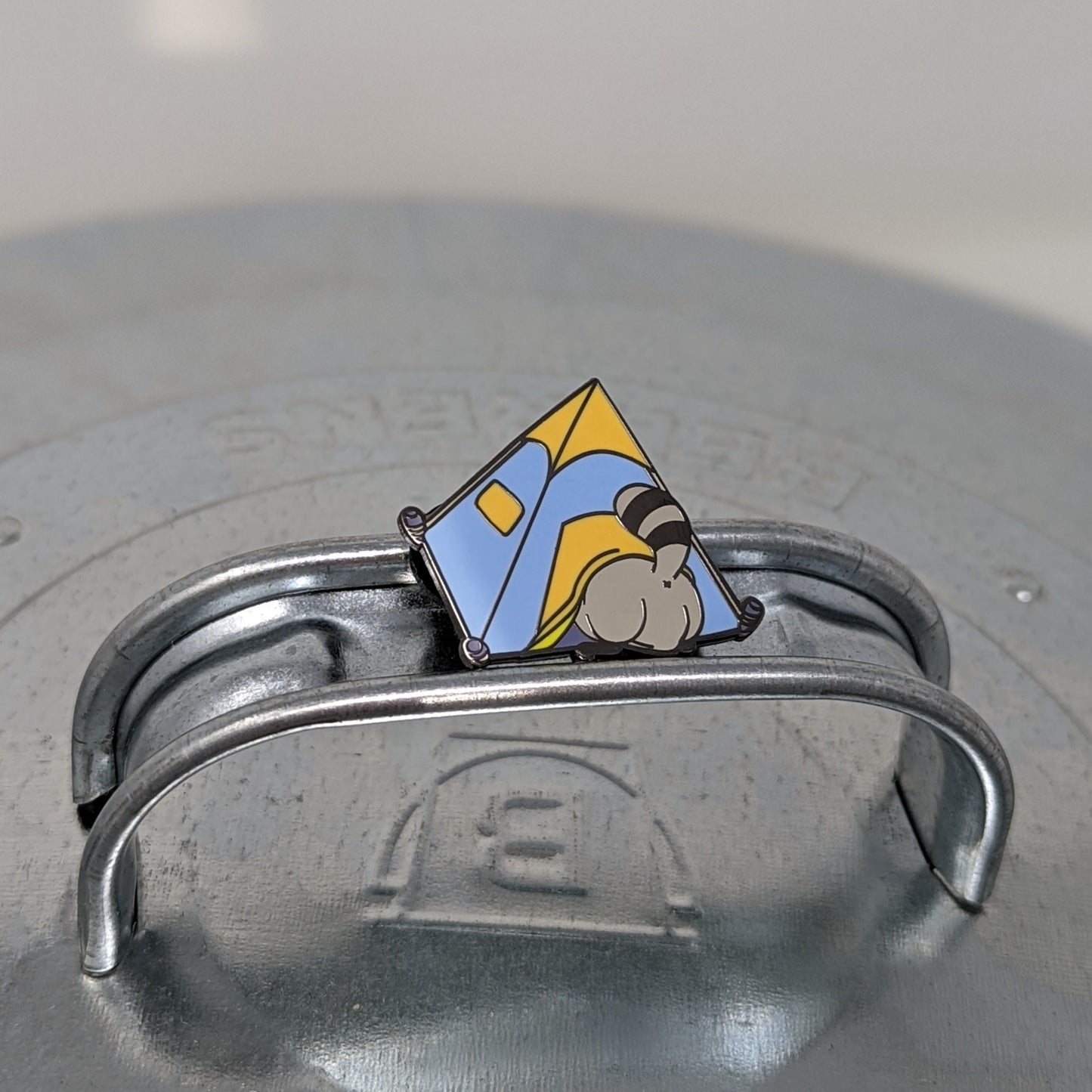 A photograph of a metal trash can lid with an enamel pin on the handle. The enamel pin is of a blue and orange tent with a raccoon butt sticking out of the door.
