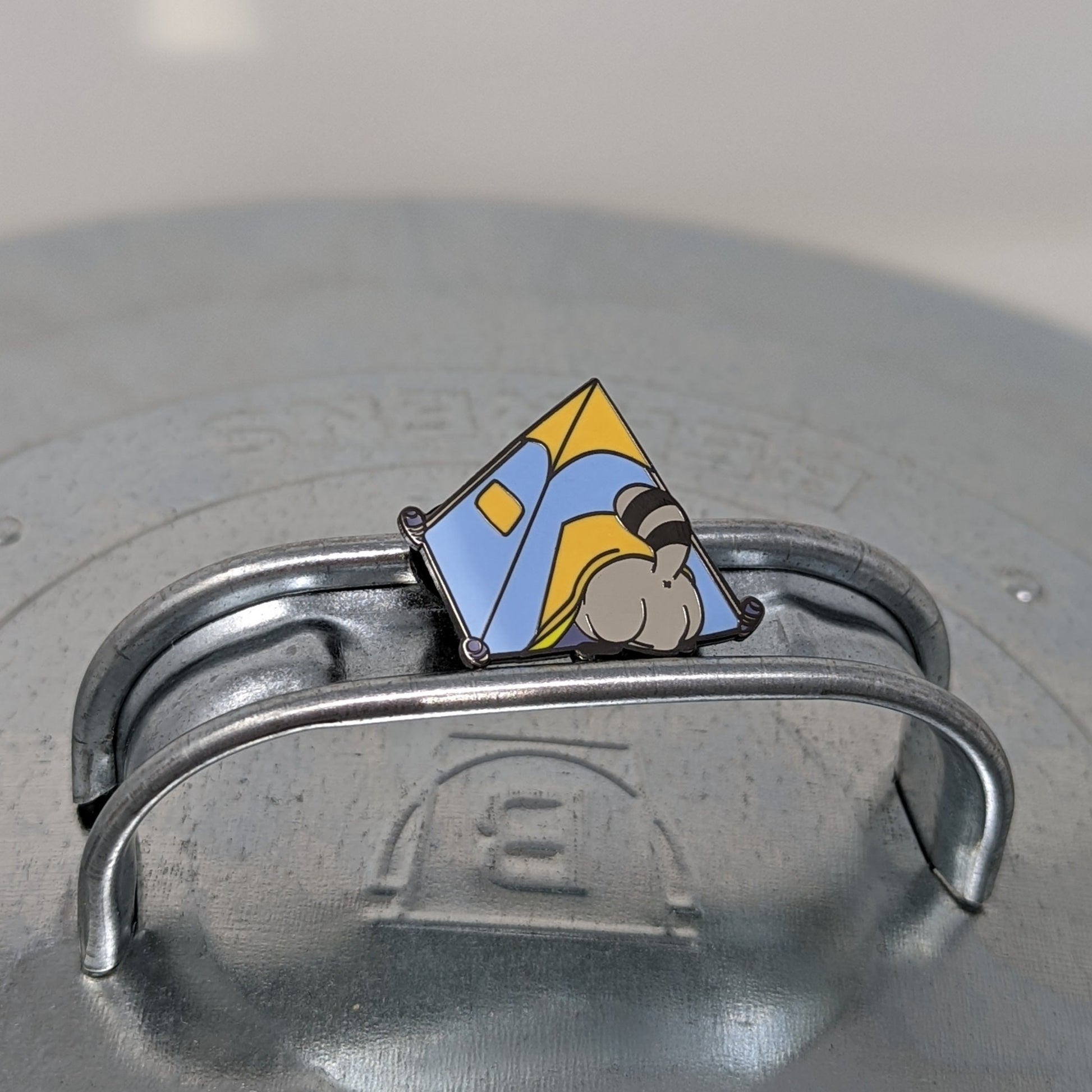 A photograph of a metal trash can lid with an enamel pin on the handle. The enamel pin is of a blue and orange tent with a raccoon butt sticking out of the door.