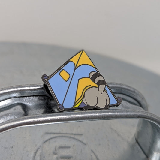 A photograph of a metal trash can lid with an enamel pin on the handle. The enamel pin is of a blue and orange tent with a raccoon butt sticking out of the door.