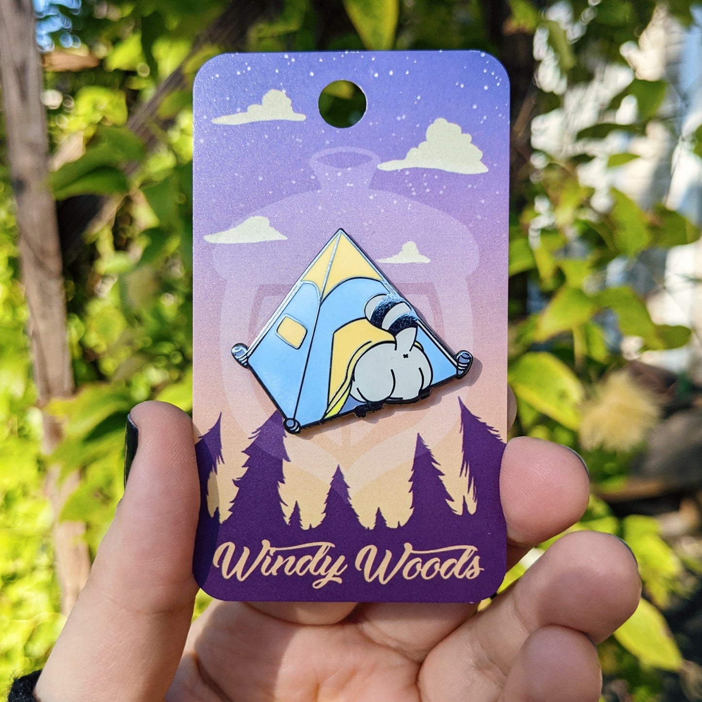 A photograph of someone holding a pin on a backing card near some flowers. The enamel pin is of a raccoon breaking into a tent. The tent is blue and orange.